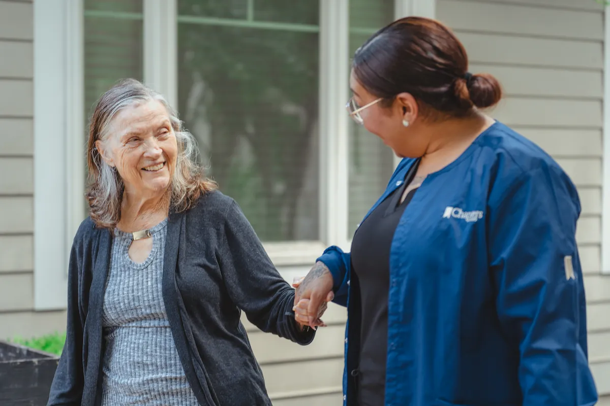 An elderly woman smiles while holding hands with a caregiver in blue scrubs outside a house, both appearing happy and engaged in conversation.