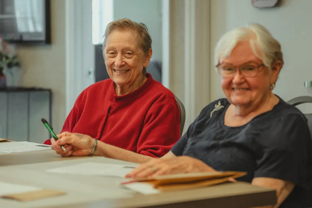 Two older women sit at a table, smiling warmly. One wears a red sweater and holds a pen, appearing to write on papers in front of them. Both seem engaged and happy, seated in a bright indoor setting.